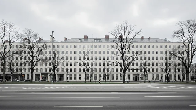 Ein langgestreckter heller Verwaltungsbau der Nachkriegszeit an einer breiten leeren Stadtstraße unter grauem Himmel, davor kahle Bäume auf einem schmalen Grünstreifen