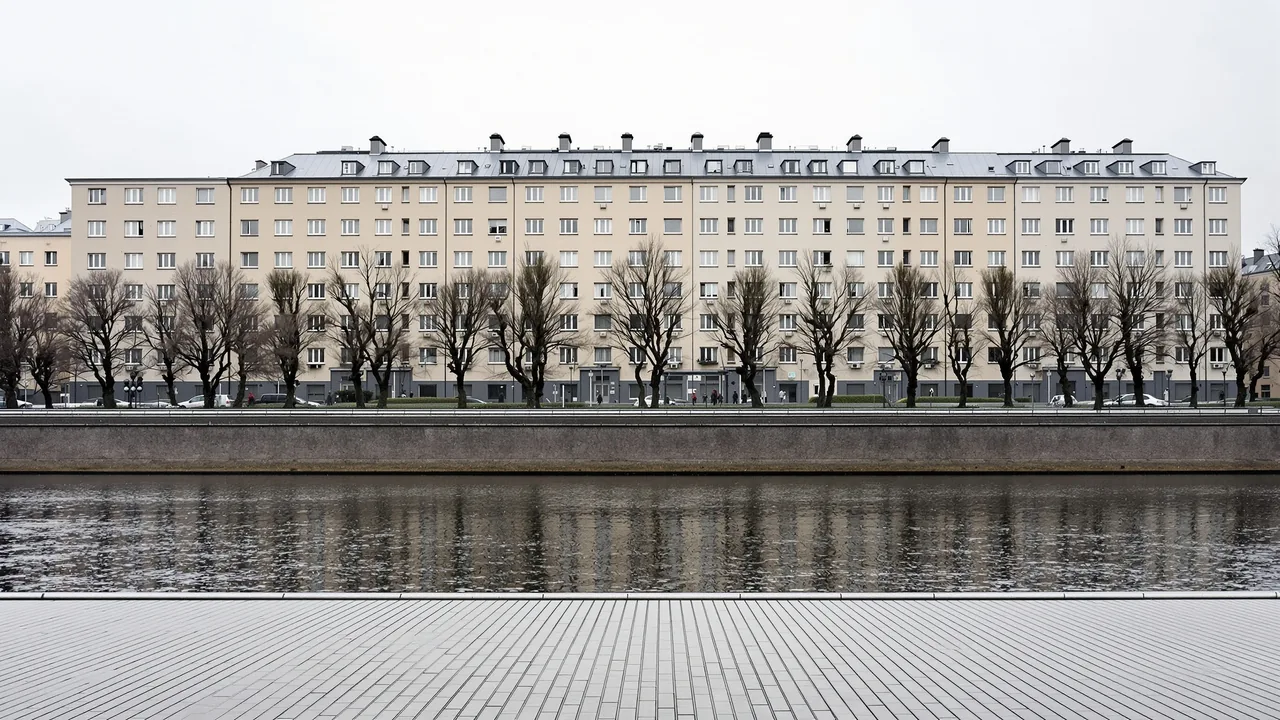 Eine graue Flusspromenade in einer deutschen Stadt im Winter, kahle Kopfplatanen vor der Reihe schlichter verputzter Nachkriegsblocks, davor stilles dunkles Wasser