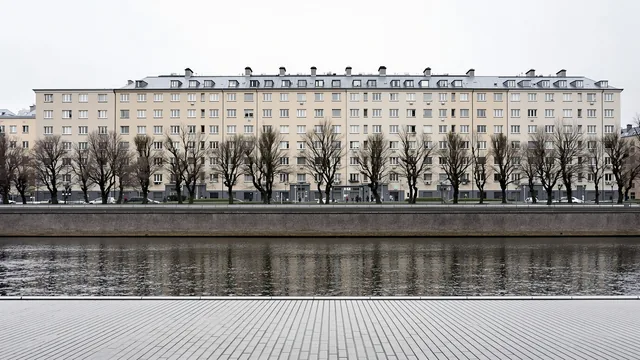 Eine graue Flusspromenade in einer deutschen Stadt im Winter, kahle Kopfplatanen vor der Reihe schlichter verputzter Nachkriegsblocks, davor stilles dunkles Wasser
