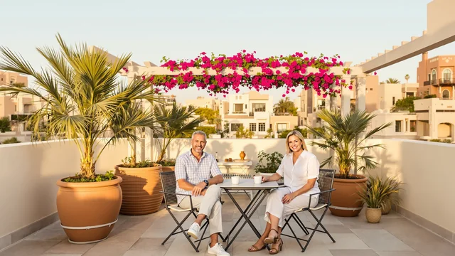Ein Mann und eine Frau sitzen am späten Nachmittag an einem kleinen Tisch mit zwei Kaffeetassen auf einer Dachterrasse in einem Wohnviertel von Dubai und lächeln in die Kamera, umgeben von Palmen in Terrakottakübeln und einer Pergola mit pinkfarbener Bougainvillea
